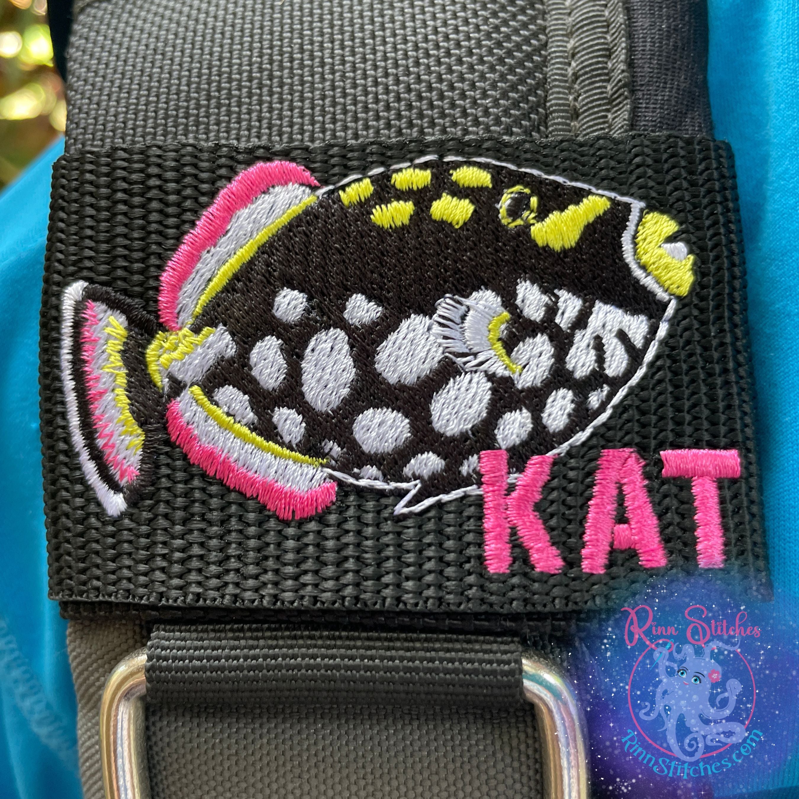 Personalized Clown Triggerfish BCD Tag with 'Kat' embroidered in Font #2 Aloha Beaches thread in Fuchsia pink, featuring black polka-dotted triggerfish design on black webbing with Velcro closure by Rinn Stitches on Maui, Hawaii