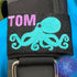 Embroidered bcd tag with a he'e (octopus) design in teal and the name tom in purple thread on the top left of the black webbing. Made by Rinn Stitches in Maui, Hawaii