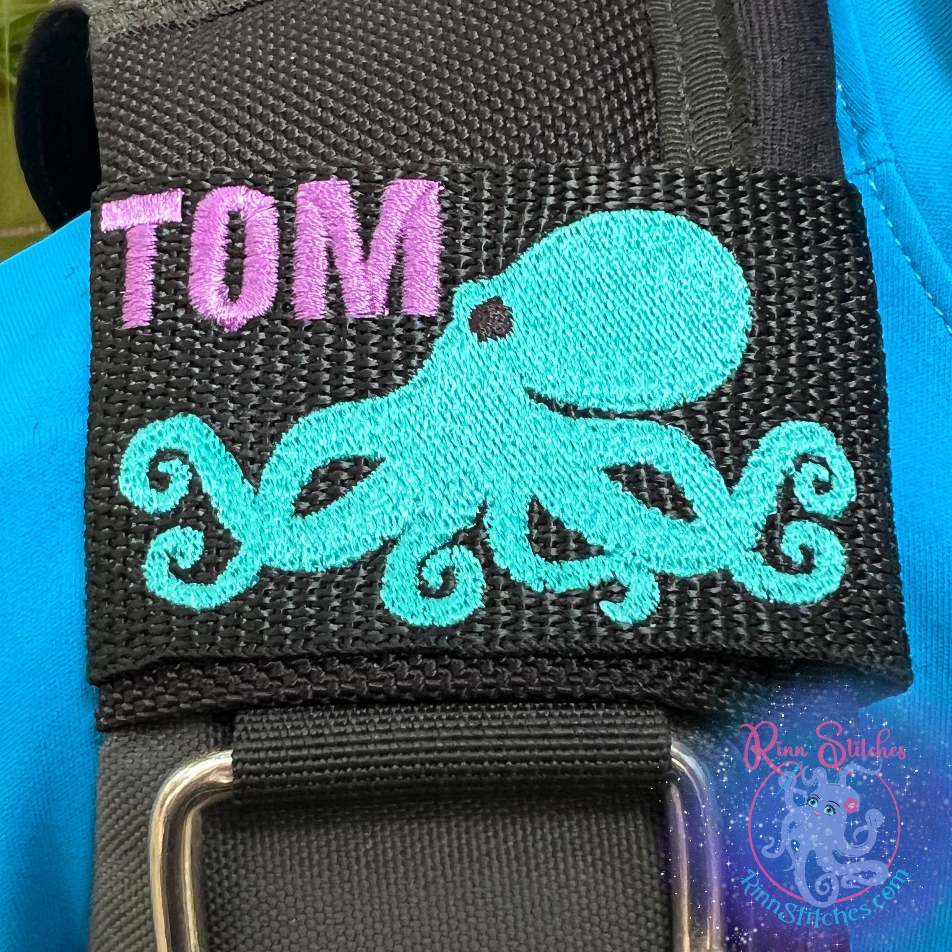 Embroidered bcd tag with a he'e (octopus) design in teal and the name tom in purple thread on the top left of the black webbing. Made by Rinn Stitches in Maui, Hawaii