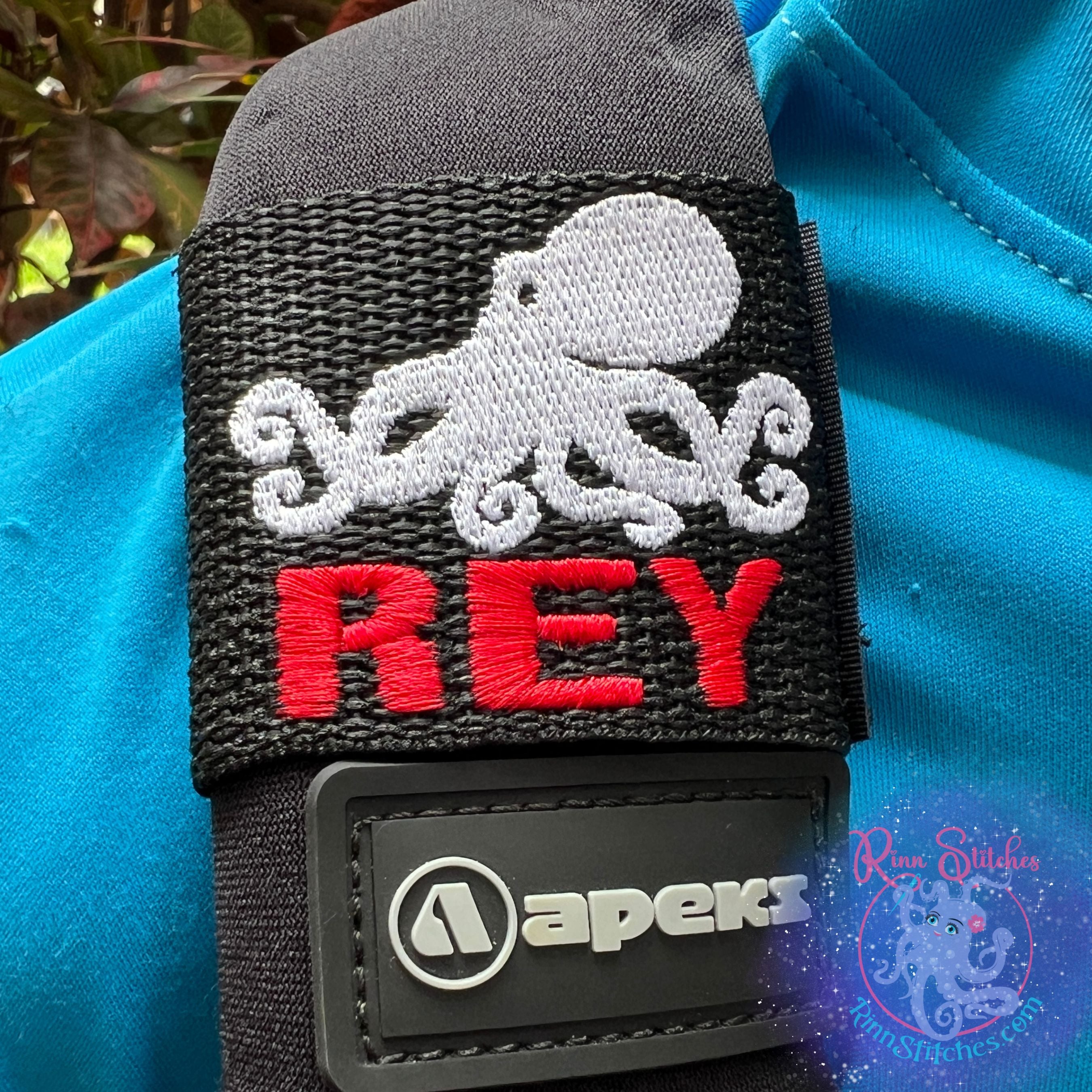 Embroidered bcd tag with a he'e (octopus) design in silver and the name Rey in red thread below the design on a 2x2 backplate and wing size tag. Made by Rinn Stitches in Maui, Hawaii